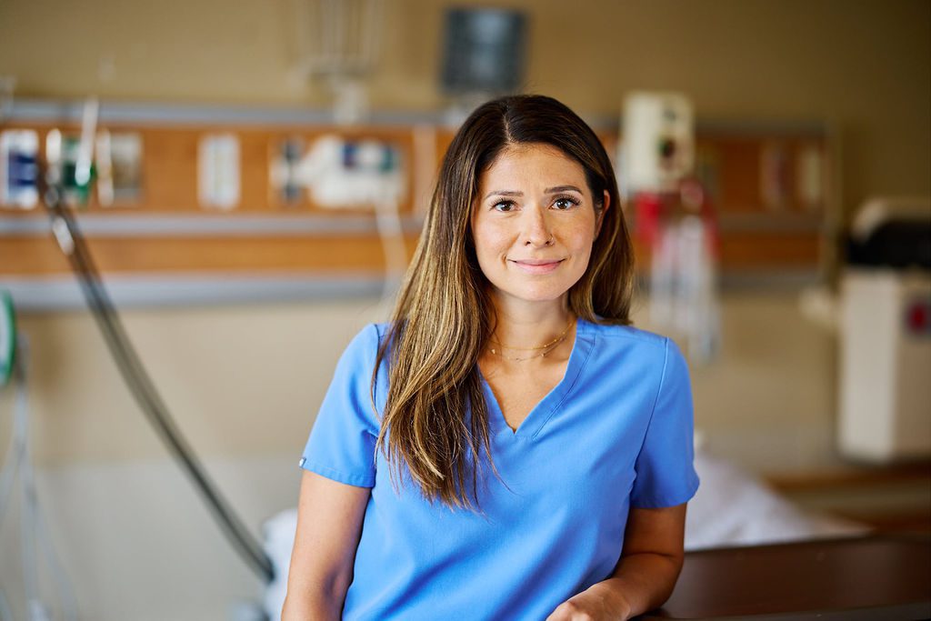A nurse in scrubs is standing next to an empty hospital bed smiling and looking at the camera.