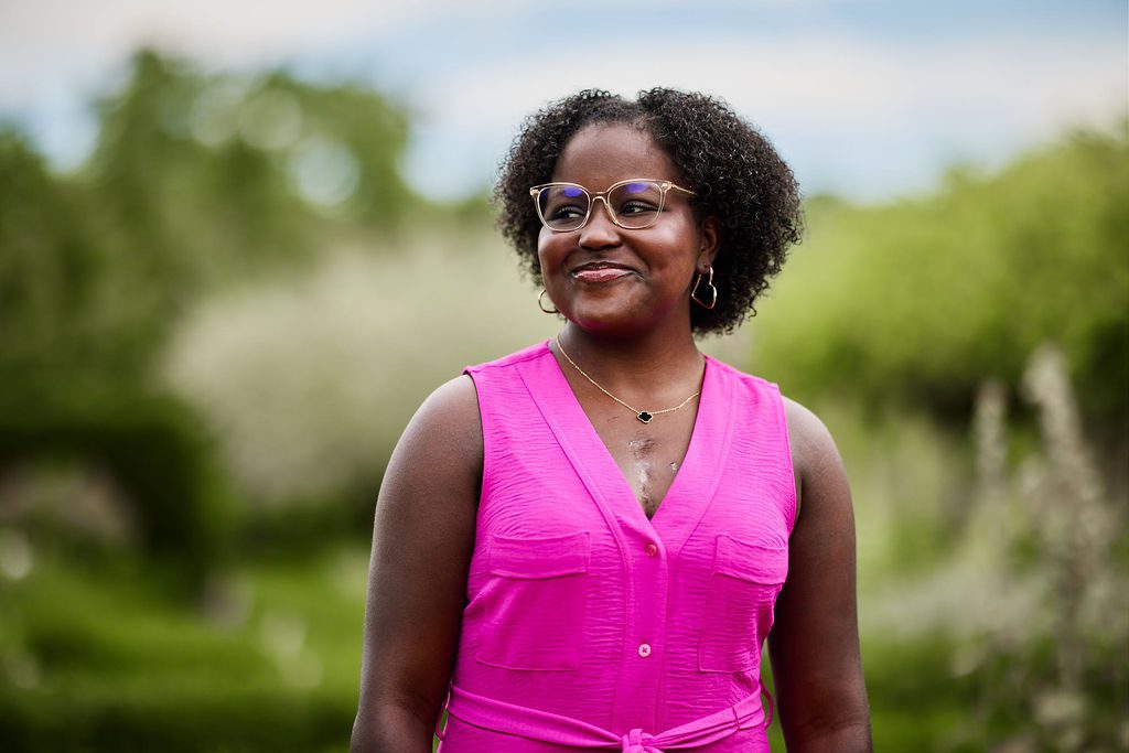 A Black woman in a bright pink dress is smiling while walking through a garden.