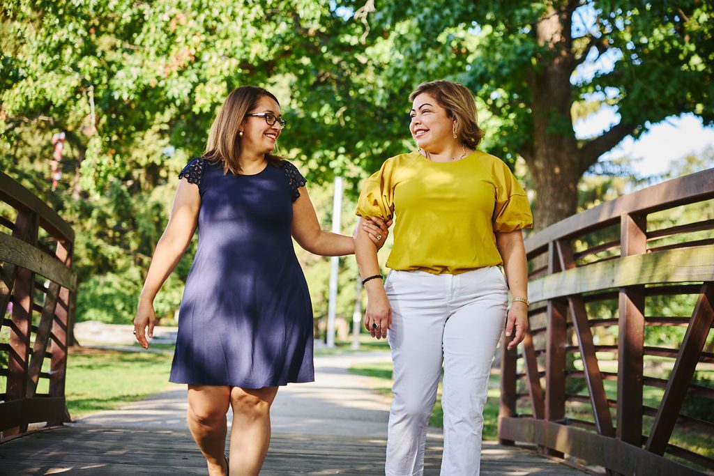 Two women are walking across a bridge in a park while chatting.