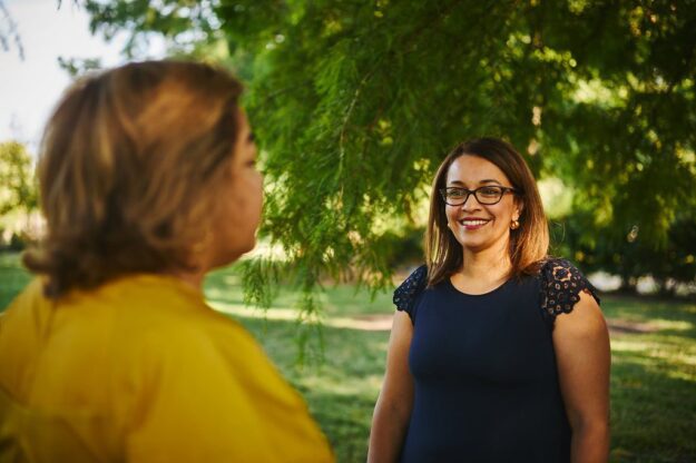 Two women looking at each other while standing in a park.